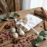 Beauty Scents wax melts with red pepper and greens, four loose on a wooden table next to rose petals, star anise, and cinnamon sticks, with a white linen bag lying on its side filled with wax melts in the background.