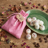 Three Beauty Scents wax melts with red pepper and greens on a rustic wooden table next to a green plate with eight more wax melts, pink linen bag, rose petals, and coffee beans.