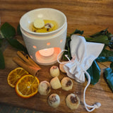 Six Beauty Scents wax melts with shredded cinnamon on a wooden table next to a white linen bag and a white oil burner melting two melts, top far angle view.