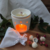 Beauty Scents wax melts with rose petals, four loose on a wooden table near tea rose buds and a white linen bag, with an oil burner in the background melting two wax melts, shown from an angle.
