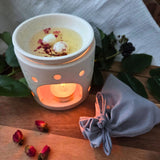 Beauty Scents wax melts with rose petals, two melts melting in an oil burner next to a grey linen bag, angled lower view.