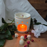 Beauty Scents wax melts with rose petals, four loose on a wooden table next to tea rose buds and a white linen bag, with an oil burner in the background melting two wax melts.