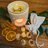 Six Beauty Scents wax melts with shredded cinnamon on a wooden table next to a white linen bag and a white oil burner melting two melts, top far angle close-up.